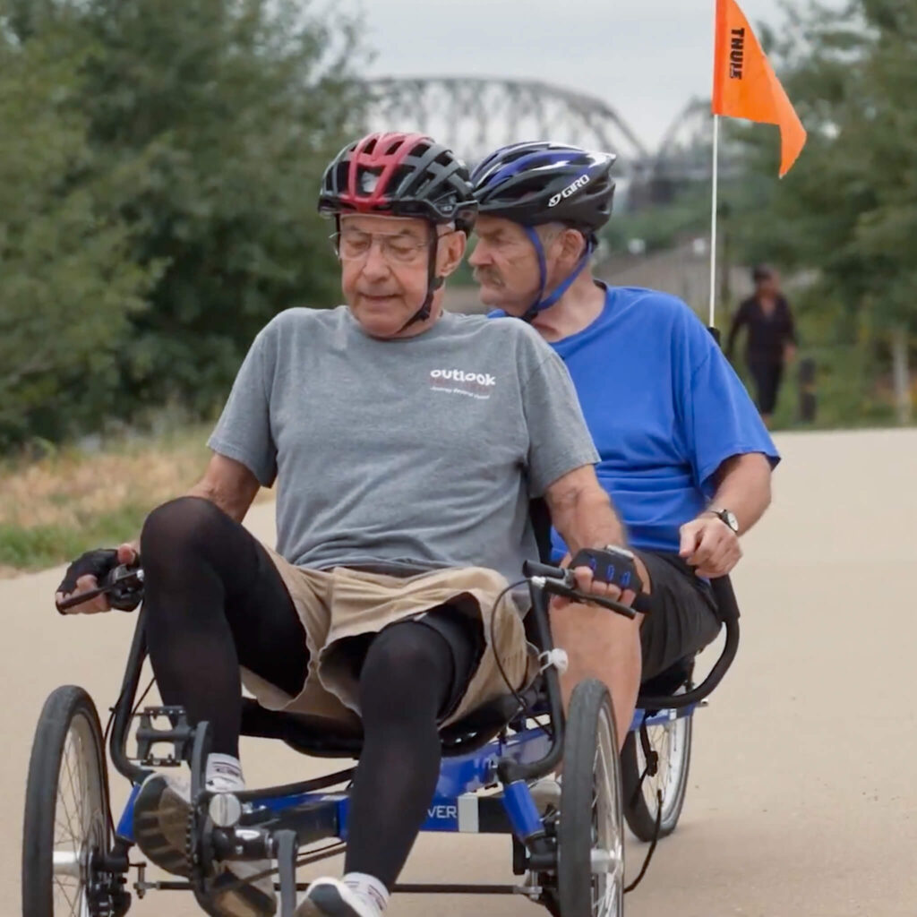 Two men wearing helmets peddle a tandem bike